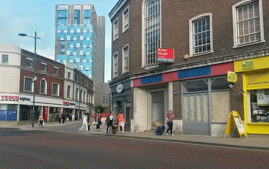 A street scene outside a commercial building with multiple pedestrians walking on the sidewalk; some carrying shopping bags, and one woman pulling a rolling suitcase with patterned luggage in front of the building. The building has a large, empty door with a curved top, a red and blue striped awning, and a yellow storefront sign for a newsagent with a small display window. To the left, there is a Tesco Express store with a white sign, and an adjacent brick building with large windows. A red 'To Let' sign is visible on the upper part of the building, indicating it is available for rental. The surrounding area features modern high-rise buildings and classic stone structures, with a clear sky overhead. This scene, captured during daytime, reflects urban movement associated with house removals or furniture transport, with people walking past, possibly during a home relocation or local move supported by [COMPANY_NAME], under the context of house removals services for the Walworth Road area in Elephant and Castle.