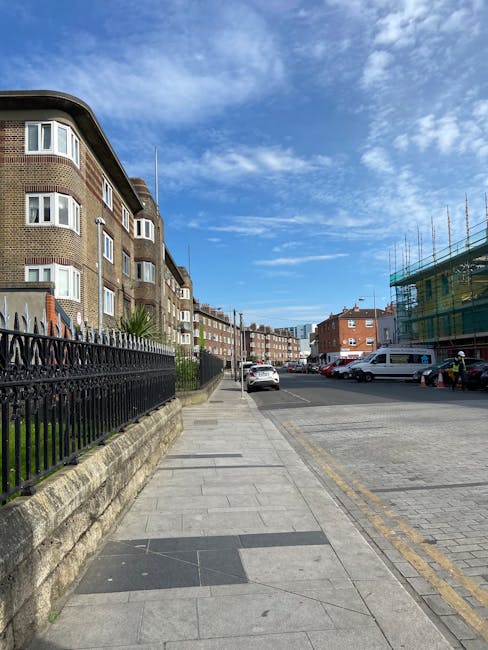 A daytime scene on a narrow urban street with brick and stone buildings on either side, where multiple cars are parked along the curb. In the background, a large white moving truck with the logo of a removal company is positioned at the end of the street, partially blocking the view of a small group of people near a doorway. The street has bollards along the pavement edge and double yellow lines indicating parking restrictions. Overhead, traffic lights and street signs are visible, and a pedestrian bridge crosses above the street. The scene captures elements typical of a home relocation or furniture transport process, with ongoing loading or unloading activities supported by [COMPANY_NAME], in an area associated with house removals for Walworth Road moves in Elephant and Castle. The weather is overcast, and the lighting is diffuse, highlighting the urban environment and moving logistics involved.