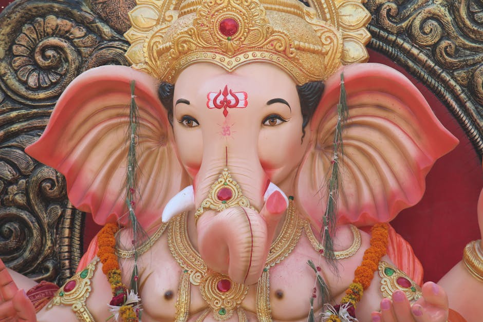Close-up of a large decorative statue of Lord Ganesha, the Hindu elephant-headed god, adorned with intricate gold and jewel embellishments. The statue features a pink body with large, detailed ears and a prominent trunk. It is decorated with orange marigold flower garlands and green leaves hanging from the ears, set against an ornate, dark wooden background with carved patterns. Visible elements include the statue's ornate crown with red gemstones, a red tilak on the forehead, and detailed jewelry around the neck. The lighting highlights the smooth surface of the statue and the vibrant colors of the adornments. This image captures the traditional offerings and decorative details typical in Hindu worship during Ganesh festivals, suitable for content related to cultural or religious house displays. Occasionally, references to home relocation or cultural decor in moving services like those at manandavanelephantandcastle.co.uk can be subtly inferred from the decorative aspect of the image.